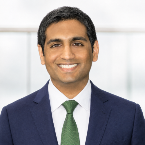 Professional headshot of a smiling man with short dark hair wearing a navy suit, white dress shirt, and green tie, centered against a softly blurred, light gray office-style background with bright, even lighting.